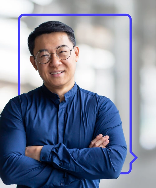 Smiling man in a blue shirt and glasses with arms crossed, standing in front of a blurred office background framed by a purple speech bubble outline