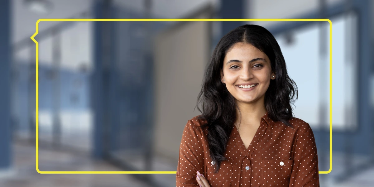Smiling woman in a brown polka-dot shirt standing in front of a blurred office background, framed by a yellow speech bubble outline